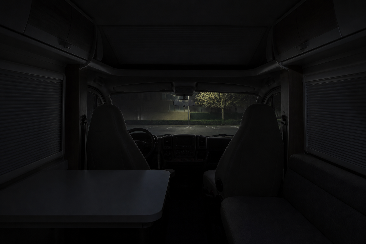 Vue de l'intérieur d'un camping-car garé sur un parking de ville belge, lumières éteintes, stores fermés, nuit tombée, sans accessoires sortis à l'extérieur